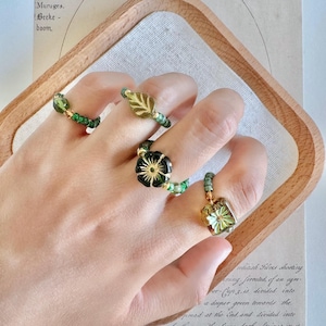 May include: Four green and gold rings with different designs. The rings are on a hand wearing a white shirt. The rings feature a leaf, a flower, and two square designs.