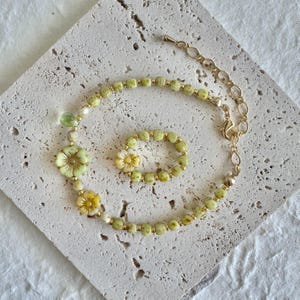 Puede incluir: Un delicado conjunto de joyas con collar y anillo. El collar y el anillo están adornados con cuentas verde claro, pequeñas cuentas doradas y detalles en forma de flor en tonos amarillos y verdes. El collar tiene una cadena dorada.