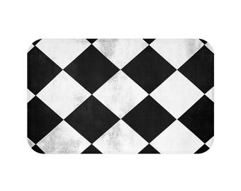 May include: Rectangular bath mat with a black and white checkered pattern. The design features alternating black and white diamond shapes, creating a classic and bold visual. The mat has a slightly distressed, textured appearance.