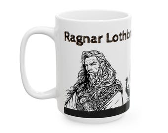 Puede incluir: Taza de cerámica blanca con una ilustración en blanco y negro de un hombre barbudo con pelo largo, y el texto "Ragnar Lothbrok" en letras marrones. La taza tiene un asa grande y una banda negra en la parte inferior.