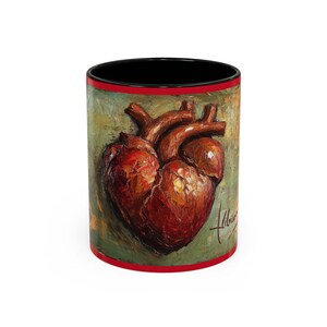 May include: A ceramic mug featuring a detailed painting of a human heart. The artwork is in shades of red and brown against a green and gold background. The mug has a black interior, a red rim, and a white exterior.