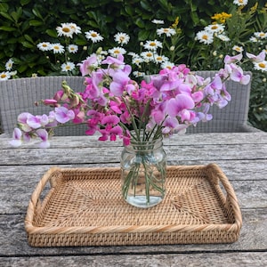 May include: A rectangular woven wicker tray with handles holds a clear glass jar filled with water and pink and purple sweet pea flowers. The tray sits on a weathered wooden table. White daisies and greenery are in the background.