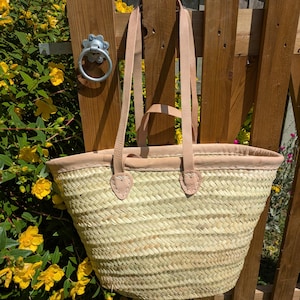 May include: A woven straw tote bag with tan leather handles and trim. The bag is hanging on a wooden fence with a metal ring latch. Yellow flowers are in the background.