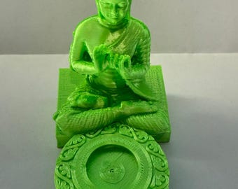 May include: A green 3D printed Buddha statue sitting on a square base with a round, decorative incense burner. The Buddha is in a meditative pose with hands clasped in front of the chest.