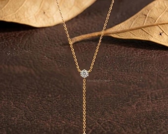 Puede incluir: Un delicado collar de oro con un colgante redondo de piedra preciosa brillante y un dije en forma de lágrima. El collar se exhibe sobre un fondo marrón con hojas secas, resaltando la elegancia de la joya.