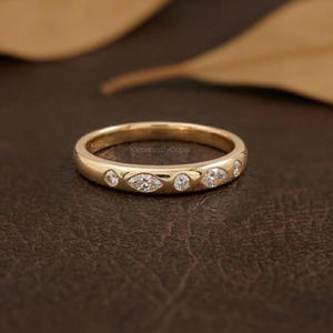 May include: A gold band ring featuring clear gemstones in round and marquise cuts. The ring is set on a dark brown surface. The text "KleodoraByGopal" is visible on the ring.