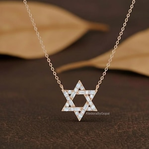 May include: A gold-colored necklace featuring a Star of David pendant. The pendant is encrusted with small, sparkling white gemstones. The necklace chain is delicate and thin. The background is a blurred mix of brown and tan.