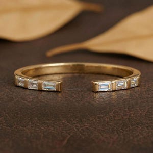 May include: A gold-coloured open ring with a series of rectangular, clear gemstones. The ring has a gap in the centre, and the gemstones are evenly spaced along the band. The ring is set against a brown background with blurred leaves.