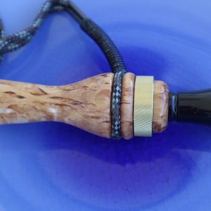 Op de afbeelding: Een eendenlokker met een houten behuizing met een lichtbruine, patroonafwerking. Het heeft een zwart mondstuk, metalen ringen en een gevlochten koord. De lokker is ontworpen voor de jacht.