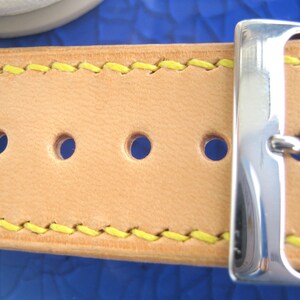 May include: Close-up of a light tan leather belt with yellow stitching. The belt has three holes and a silver buckle. The stitching and buckle provide a contrasting design element.