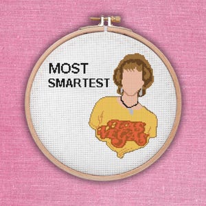 May include: Cross-stitch embroidery hoop with a yellow shirt with the text "Most Smartest" and a red and orange graphic of a person's face. The person has brown hair and is wearing a necklace.