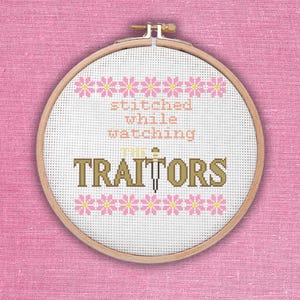 Op de afbeelding: Een borduurring met kruissteek met de tekst "stitched while watching THE TRAITORS". De tekst is in een mix van kleuren, waaronder roze, bruin en beige. Roze bloempatronen omlijsten de boven- en onderkant van de tekst. De achtergrond is wit.