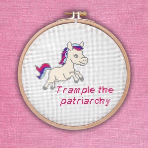 May include: A cross-stitch hoop featuring a cream-colored unicorn with a pink and blue mane and tail, jumping. The text "Trample the patriarchy" is stitched in pink below the unicorn. The hoop is set against a pink background.