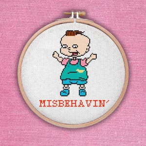 Puede incluir: Aro de bordado de punto de cruz con una imagen de dibujos animados de un bebé con un atuendo azul y rosa. El texto "MISBEHAVIN'" está debajo de la imagen.