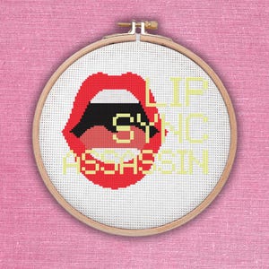 May include: A white embroidery hoop with a red open mouth with a black tongue and the words "Lip Sync Assassin" in yellow.