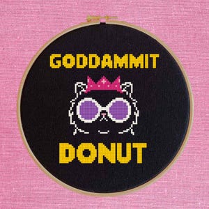 May include: A circular wooden embroidery hoop containing black fabric with a cross-stitch design. The design features a white-outlined cat wearing a pink crown and purple sunglasses, with the yellow text GODDAMMIT above and DONUT below.