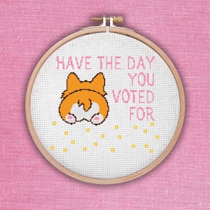 May include: A cross-stitch embroidery hoop with the text "HAVE THE DAY YOU VOTED FOR" in pink. The design features a cartoon corgi butt in orange and white, with yellow star accents. The hoop is set against a pink fabric background.