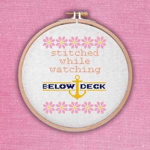 May include: A cross-stitch embroidery hoop with the words "stitched while watching" above the words "BELOW DECK" and an anchor. Pink flowers are stitched above and below the text. The background is white, and the hoop is set against a pink background.