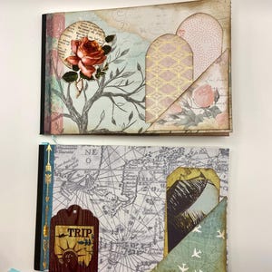 May include: Two handmade scrapbook albums. The top album features a rose and tree design with gold accents. The bottom album has a world map design with a "TRIP" tag and a blue ribbon. Both albums have decorative pockets.