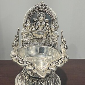 Puede incluir: Diya de plata con detalles intrincados, que presenta una figura de diosa sentada con una base de flor de loto. El diya es una l&aacute;mpara de aceite tradicional india que se utiliza para ceremonias religiosas y festivales.