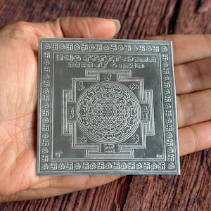 May include: A silver square with intricate geometric patterns and text. The design includes a central circular motif and a border of repeating symbols. The object is held in a hand against a wooden background.