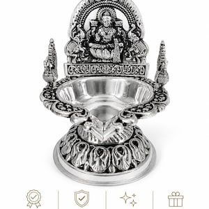 Op de afbeelding: Een zilverkleurige, sierlijke olielamp met een gedetailleerd ontwerp. De lamp heeft een centrale kom, een decoratieve basis en een gedetailleerde boog met een zittende figuur en olifantenmotieven. De woorden "Premium Quality", "Durable & Long Lasting" en "Elegant Design" zijn zichtbaar.