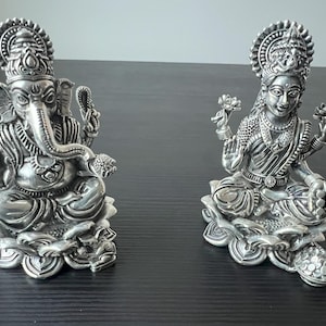Op de afbeelding: Twee zilveren beelden van hindoe&iuml;stische goden, Ganesha en Lakshmi, zittend op lotusbloem bases. De beelden zijn ingewikkeld gedetailleerd en hebben een gepolijste afwerking.