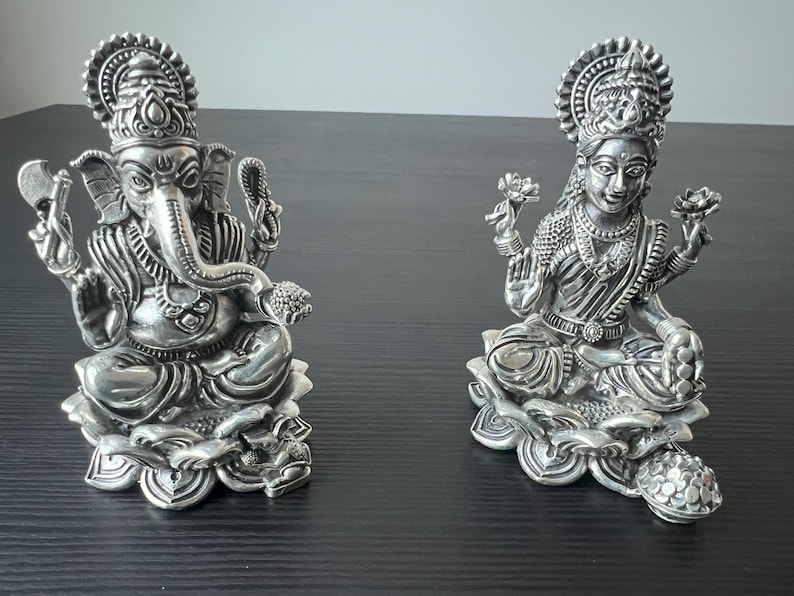 Op de afbeelding: Twee zilveren beelden van hindoe&iuml;stische goden, Ganesha en Lakshmi, zittend op lotusbloem bases. De beelden zijn ingewikkeld gedetailleerd en hebben een gepolijste afwerking.