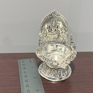 Puede incluir: Diya de plata con un dise&ntilde;o detallado que presenta una deidad sentada en un trono. La diya mide aproximadamente 7,5 cm de alto.