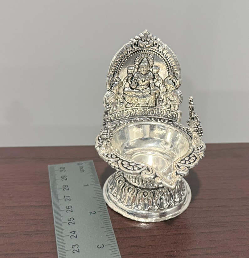 Puede incluir: Diya de plata con un dise&ntilde;o detallado que presenta una deidad sentada en un trono. La diya mide aproximadamente 7,5 cm de alto.
