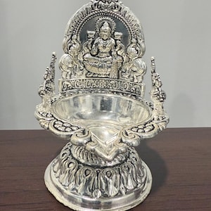 Puede incluir: Una l&aacute;mpara Kamakshi de plata con detalles intrincados, que pesa 84 gramos y est&aacute; marcada con BIS 925. La l&aacute;mpara presenta una figura de deidad sentada en un trono de loto.