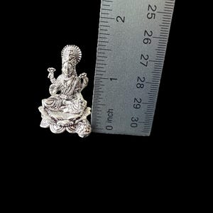 Op de afbeelding: Zilveren Lakshmi-godinbeeldje dat op een lotusbloem zit. Het beeldje is ongeveer 2,5 cm hoog en wordt naast een liniaal getoond voor schaal.