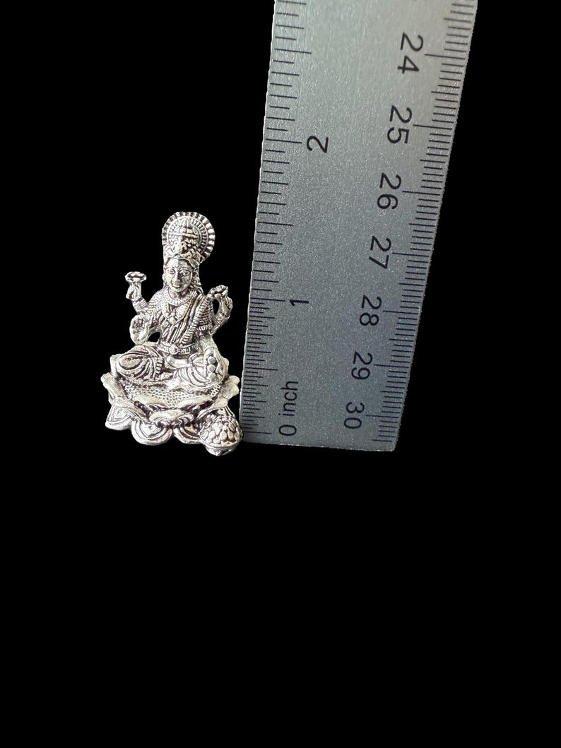 Op de afbeelding: Zilveren Lakshmi-godinbeeldje dat op een lotusbloem zit. Het beeldje is ongeveer 2,5 cm hoog en wordt naast een liniaal getoond voor schaal.