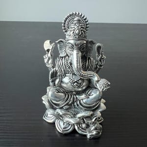 Op de afbeelding: Een zilveren Ganesha-beeld dat op een lotusbloembasis zit. Het beeld is ingewikkeld gedetailleerd en heeft een traditioneel ontwerp.