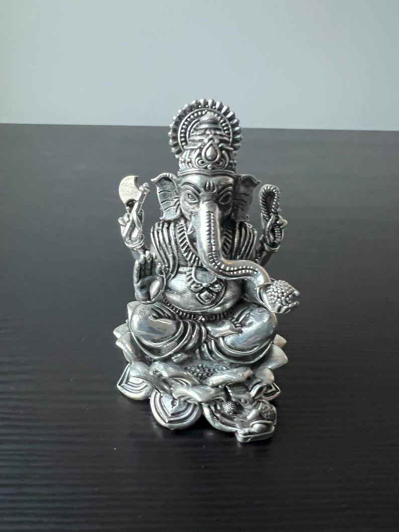 Op de afbeelding: Een zilveren Ganesha-beeld dat op een lotusbloembasis zit. Het beeld is ingewikkeld gedetailleerd en heeft een traditioneel ontwerp.