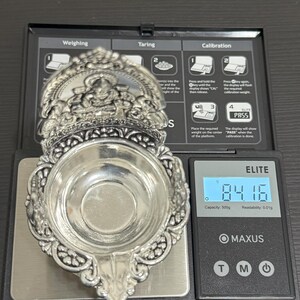 Puede incluir: Un diya de plata, una l&aacute;mpara de aceite tradicional india, se est&aacute; pesando en una b&aacute;scula digital. La b&aacute;scula marca 84,16 gramos. El diya tiene intrincados dise&ntilde;os florales y un cuenco central para aceite.