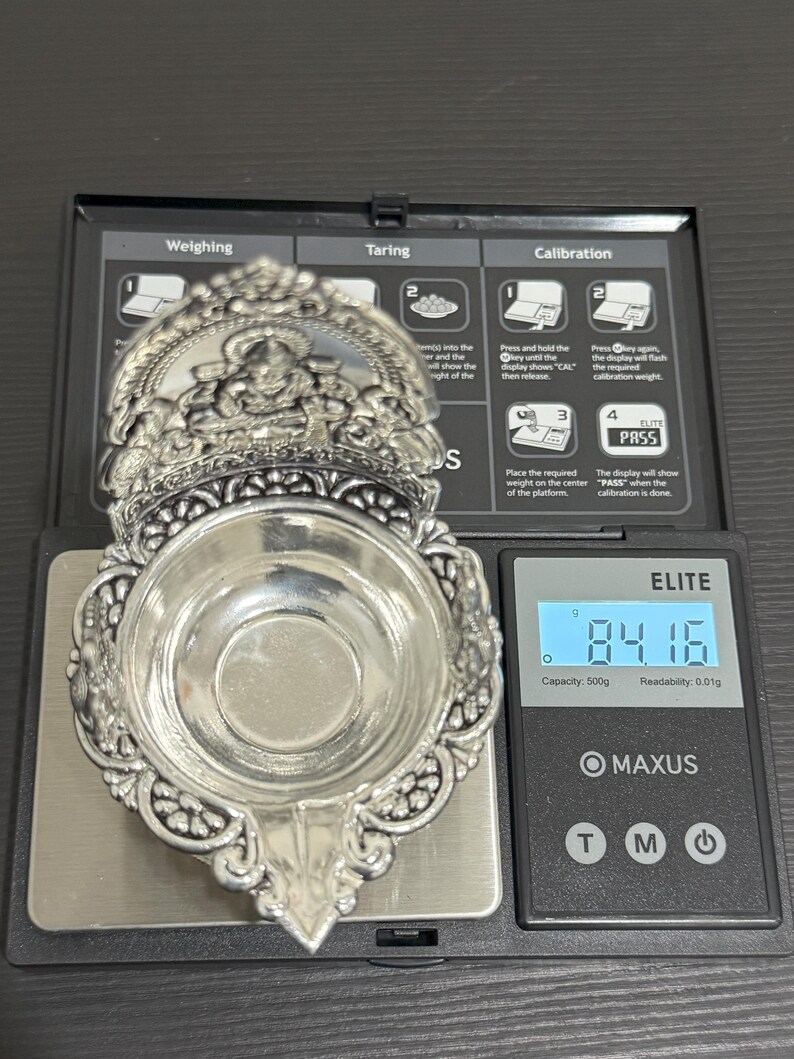 Puede incluir: Un diya de plata, una l&aacute;mpara de aceite tradicional india, se est&aacute; pesando en una b&aacute;scula digital. La b&aacute;scula marca 84,16 gramos. El diya tiene intrincados dise&ntilde;os florales y un cuenco central para aceite.