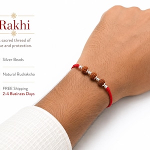 May include: A red thread bracelet featuring three brown, textured natural Rudraksha beads interspersed with small, smooth silver-colored beads. The text on the image reads Rakhi, A sacred thread of love and protection, Silver Beads, and Natural Rudraksha.