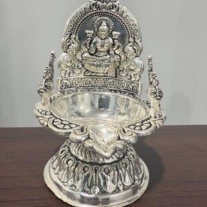 Puede incluir: Un diya de plata con detalles intrincados, que presenta una deidad sentada en un trono. El diya tiene una base redonda y un borde decorativo.