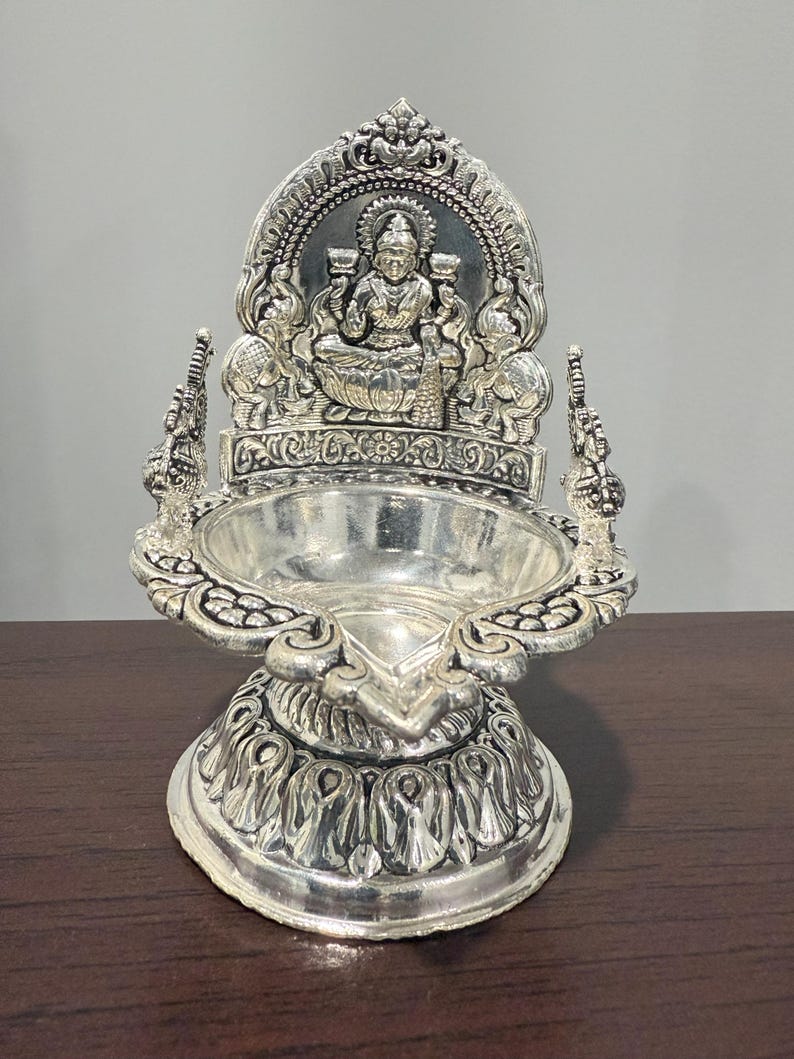 Puede incluir: Un diya de plata con detalles intrincados, que presenta una deidad sentada en un trono. El diya tiene una base redonda y un borde decorativo.