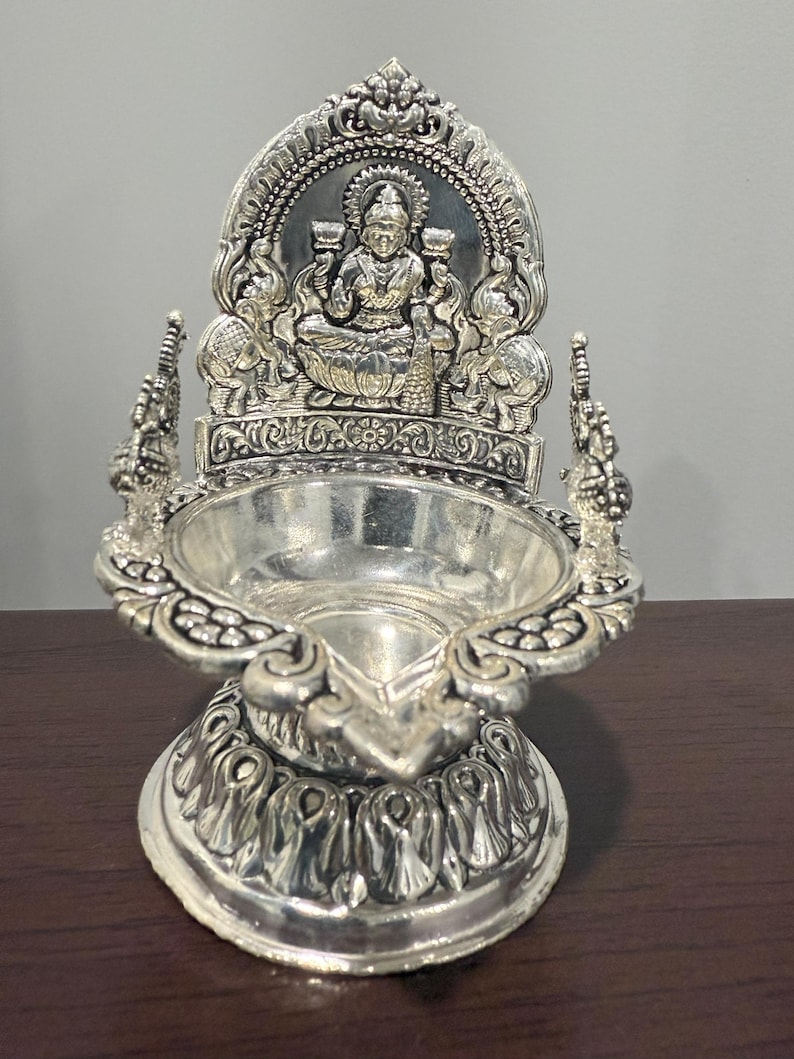 Puede incluir: Diya de plata con detalles intrincados, que presenta una figura de diosa sentada con una base de flor de loto. El diya es una l&aacute;mpara de aceite tradicional india que se utiliza para ceremonias religiosas y festivales.
