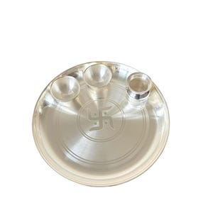 Op de afbeelding: Een zilveren puja thali set met een centraal swastika ontwerp. De set bevat een grote ronde plaat, twee kleine kommen en een cilindrische container, allemaal met een gepolijste afwerking. De items zijn op een witte achtergrond geplaatst.