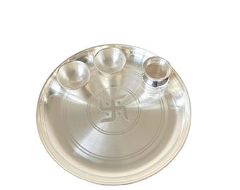 May include: A silver puja thali set with a central swastika design. The set includes a large round plate, two small bowls, and a cylindrical container, all with a polished finish. The items are arranged on a white background.