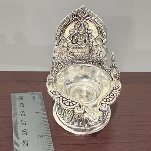 Puede incluir: Diya de plata con un dise&ntilde;o detallado que presenta una deidad sentada. La diya mide aproximadamente 5 cm de alto.