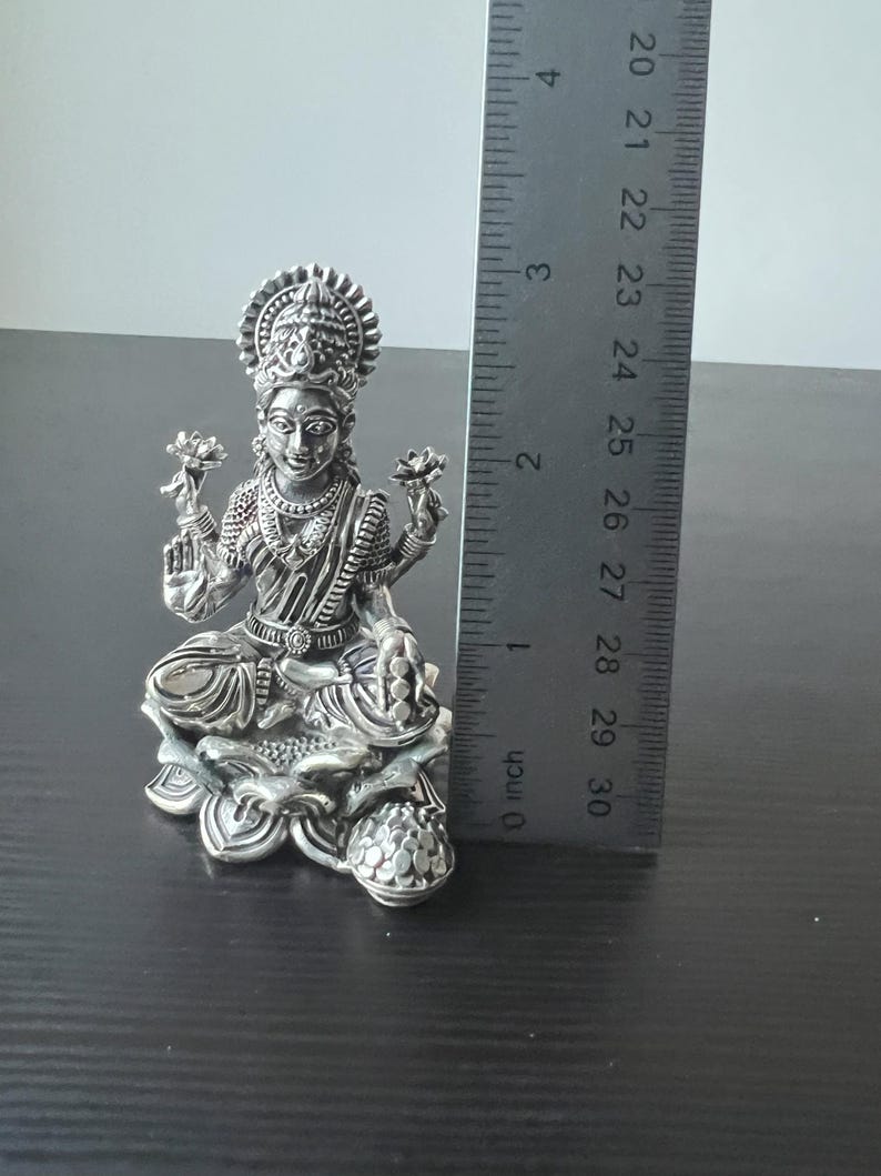 Op de afbeelding: Zilveren beeld van de hindoegodin Lakshmi zittend op een lotusbloem. Ze houdt een lotusbloem in elke hand en draagt een kroon op haar hoofd. Het beeld is ongeveer 15 cm hoog.