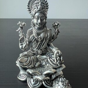 Op de afbeelding: Een zilveren beeld van de hindoegodin Lakshmi, zittend op een lotusbloem. Ze houdt een lotusbloem in elke hand en heeft een serene uitdrukking op haar gezicht.