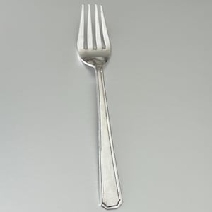 May include: A silver fork with four tines and a long, rectangular handle. The handle has a geometric design. The fork is made of a shiny, reflective metal and is set against a grey background.