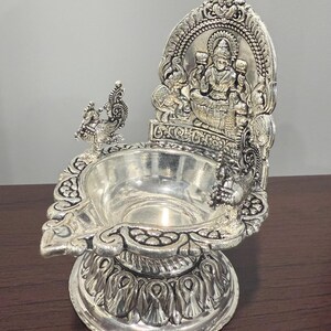 Puede incluir: Diya de plata con un dise&ntilde;o detallado que presenta una figura de diosa sentada y pavos reales. El diya tiene una base redonda y una parte superior decorativa.