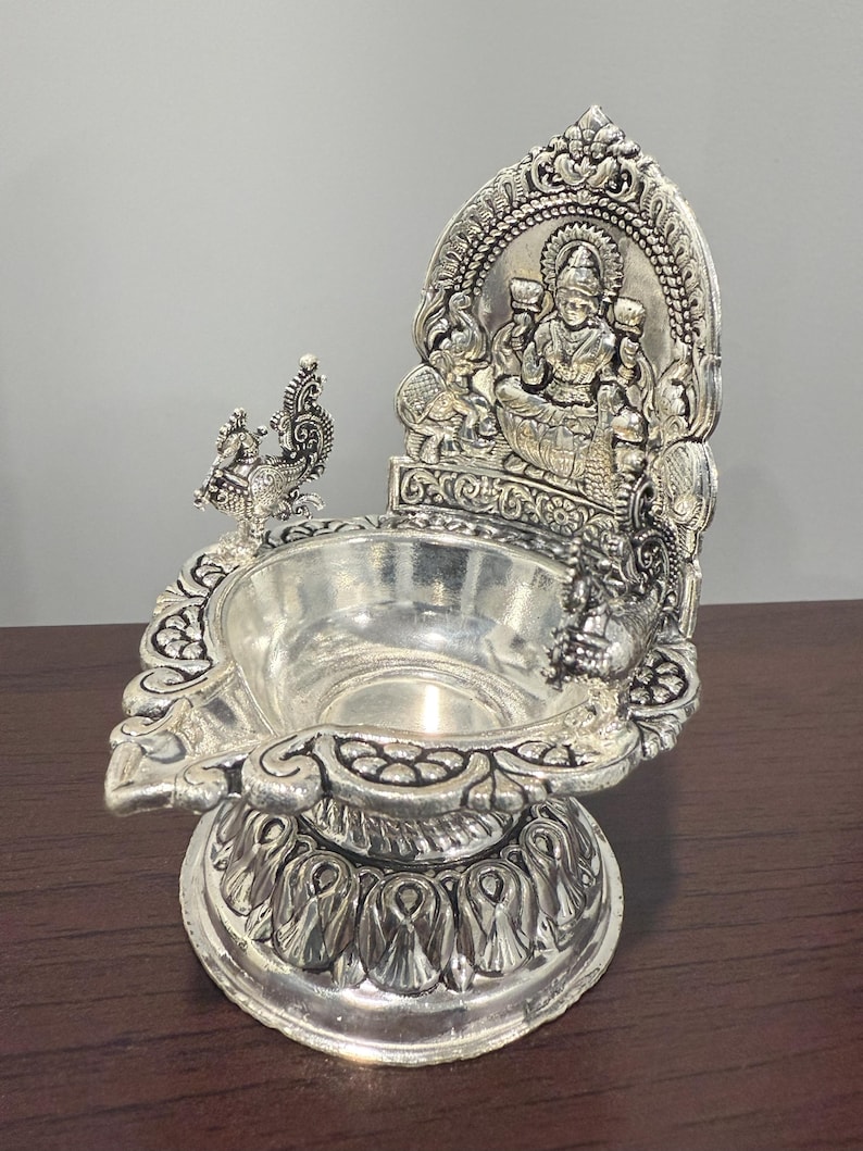 Puede incluir: Diya de plata con un dise&ntilde;o detallado que presenta una figura de diosa sentada y pavos reales. El diya tiene una base redonda y una parte superior decorativa.