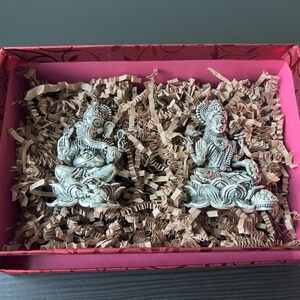 Op de afbeelding: Twee zilveren beeldjes van hindoe&iuml;stische goden, Ganesha en Lakshmi, worden getoond in een rode geschenkdoos met bruin gescheurd papier. De beeldjes zitten op lotusbloem bases.